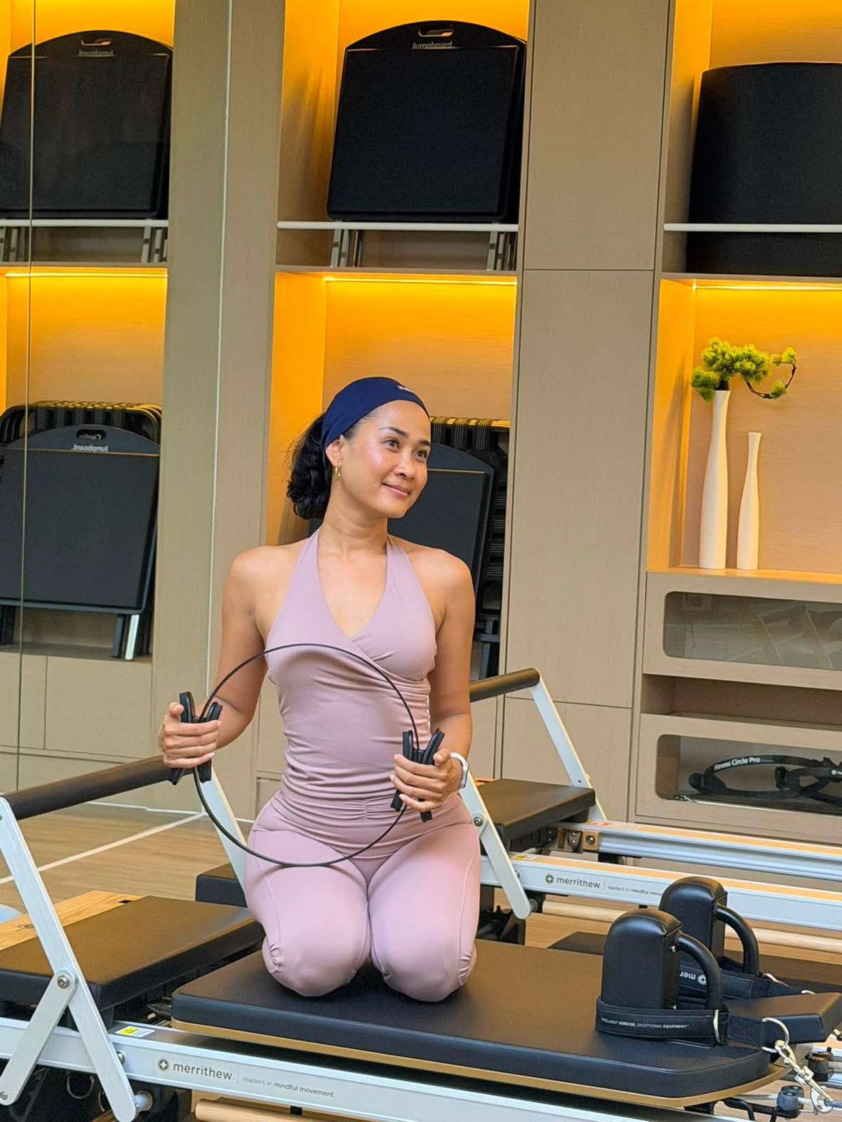 Pisey kneeling on the reformer with a Pilates ring, warm cabinet light behind her